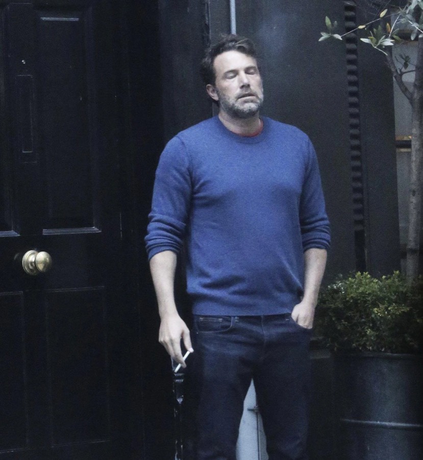 A man standing outside a black door with his eyes shut and a cigarette in one hand